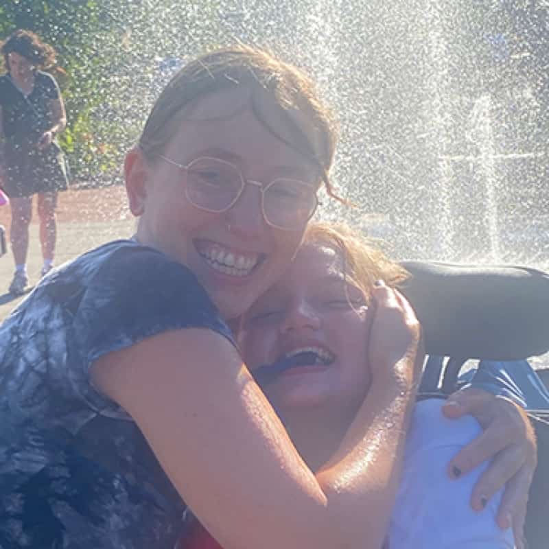 Apex-Social-Fachkraft umarmt lachend ihr Gastkind an einem Wasserspielplatz